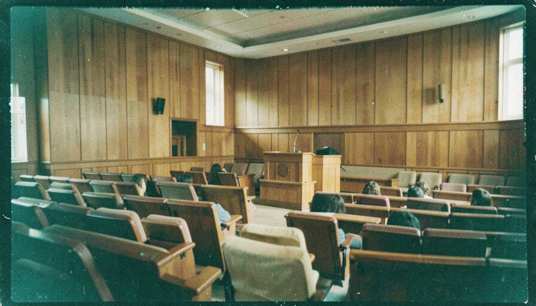 A grainy analog film photograph of a quiet university lecture hall with warm wooden paneling, rows of empty seats, and soft daylight falling across a podium — muted browns, creams, and deep shadows.